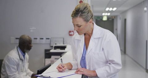 Female doctor writing patient notes at hospital reception desk with colleague in white coat