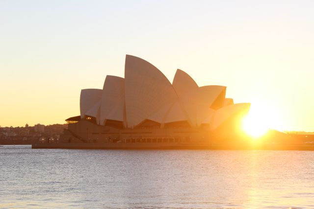 This photo captures the Sydney Opera House during sunset with a golden glow, creating a dramatic silhouette against the water. This is an iconic view of Australia's most famous landmark, making it perfect for use in travel brochures, tourism promotional materials, architecture features, and as a symbolic representation of Sydney.