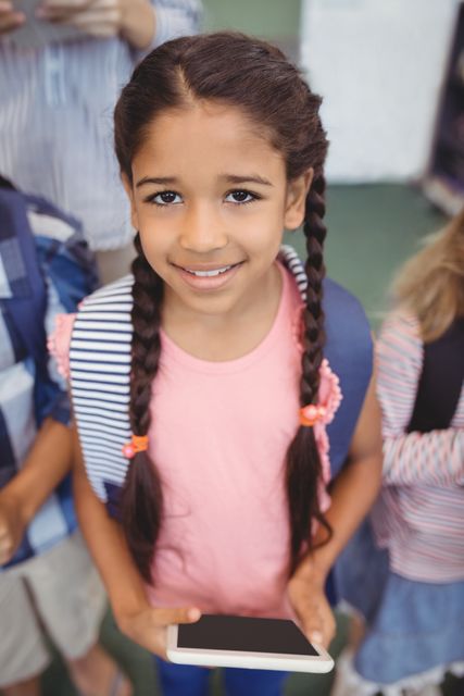 Young girl with braided hair smiling while holding a digital tablet and wearing a backpack. Ideal for educational content, back-to-school promotions, technology in education, and childhood learning materials.