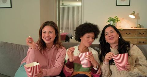Diverse women laughing and sharing popcorn on cozy gray couch at home