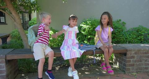 Elementary classmates sharing playful moment on school bench with backpacks and greenery