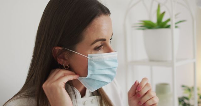 This image depicts a woman adjusting a surgical mask on her face. The close-up shot highlights health and safety precautions during the COVID-19 pandemic. Could be used in articles about safety measures, healthcare guidelines, medical advice, or public service announcements regarding the importance of wearing masks.
