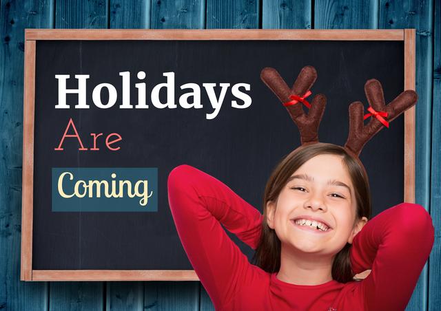 Smiling girl wearing reindeer antlers stands in front of a blackboard that reads 'Holidays Are Coming'. Useful for advertisements, holiday campaigns, festive greeting cards, and seasonal promotions. Captures the excitement and joy of the holiday season.