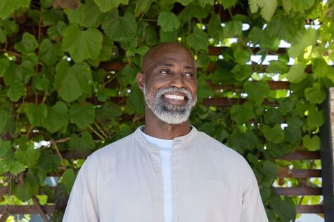 Mature african american man smiling in lush garden by wooden trellis wearing beige shirt