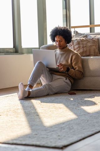 African american man working on laptop at home leaning against sofa in sunlit interior