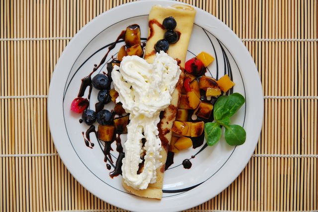 Crepe garnished with whipped cream and vibrant mix of blueberries and other fresh fruits on white plate. Ideal for use in food blogs, culinary websites, gourmet magazines, or advertisements for confectioneries and restaurants focusing on desserts and fine dining.