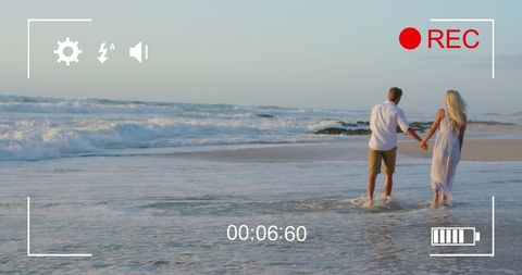 Young couple holding hands on ocean shoreline being recorded
