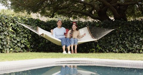 Female friends laughing on hammock by pool holding drinks and smartphone, backyard retreat