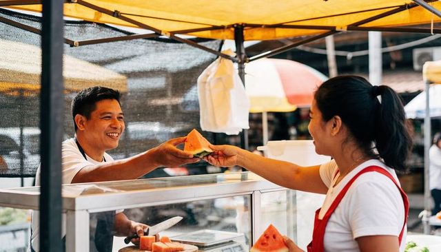 Smiling vendor handing fresh watermelon slice to customer at bustling street market