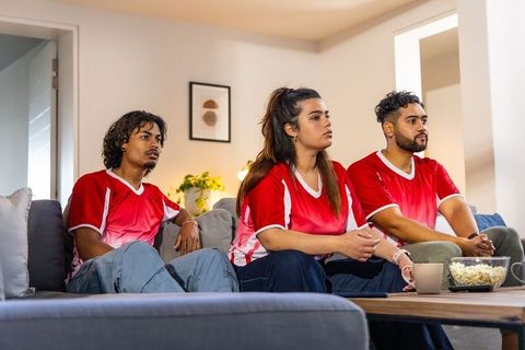 Diverse young fans wearing red jerseys watching football match nervously on sofa