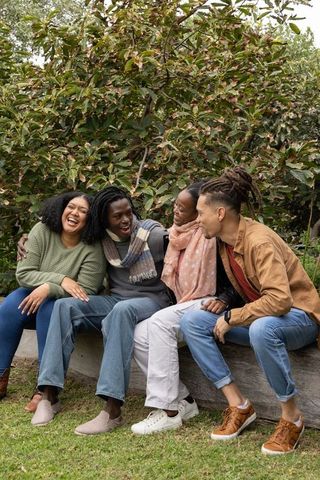 African american friends laughing on bench in autumn park wearing cozy sweaters