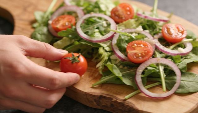 Hand placing cherry tomato on rustic cutting board with fresh greens, feta and red onion