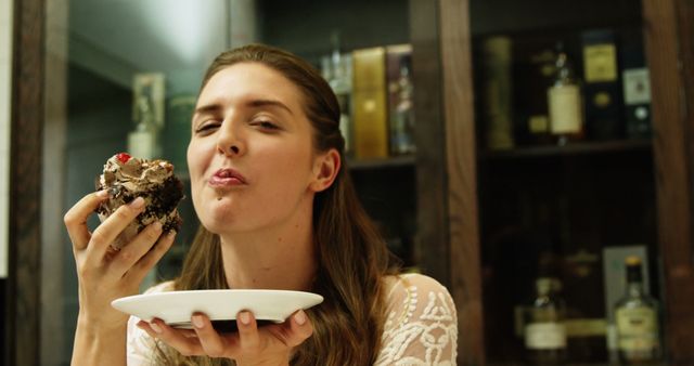 Woman is savoring a dessert, holding a white plate with the treat in one hand. Her expression denotes delight and satisfaction. Ideal for concepts related to gourmet food, joyful moments, relaxing at home, and lifestyle content.