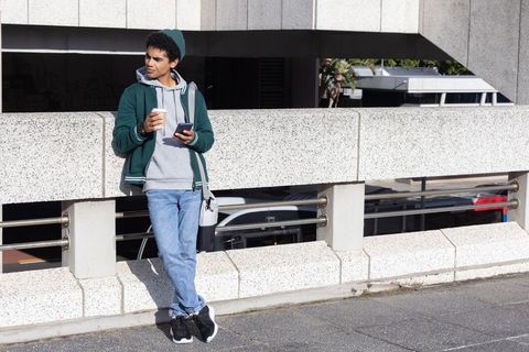 African american commuter leaning on overpass holding coffee and smartphone