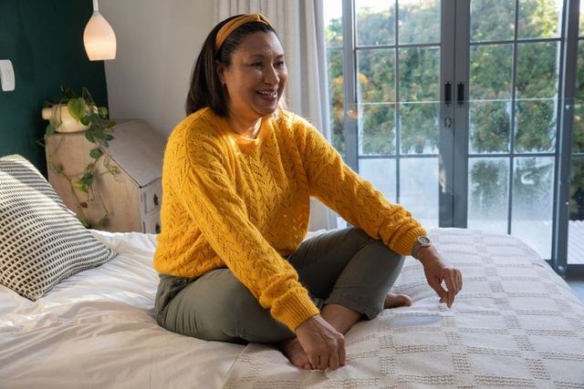 Mature woman sitting cross-legged on bed smiling in sunlit bedroom wearing yellow sweater