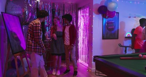 Asian woman chatting with friends at retro neon house party with disco ball and pool table