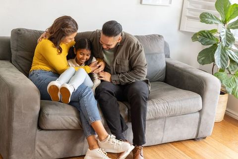 Parents cuddling daughter on cozy gray sofa in sunlit living room for family lifestyle