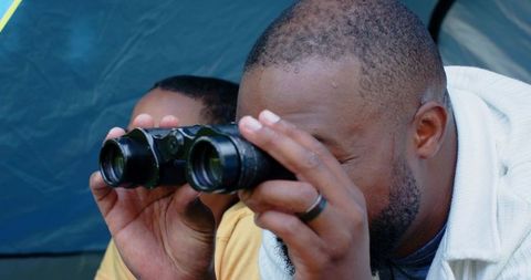 African american father guiding son with binoculars inside tent during camping trip