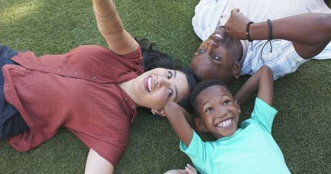 Smiling mixed-race family lying on lawn taking selfie, joyful outdoor moment