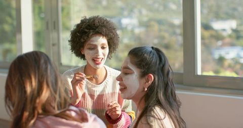 Friends applying face masks in sunlit room enjoying skincare party and self-care