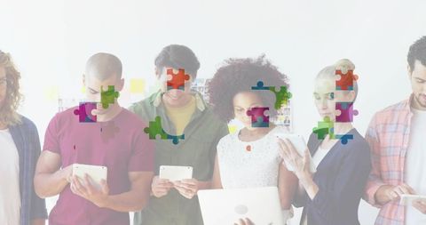 Diverse team standing using tablets and laptop with colorful jigsaw overlay