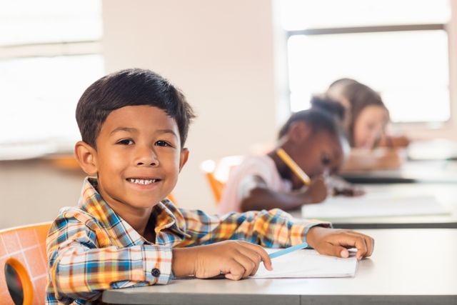 Young boy smiling while sitting at his desk in a classroom. Ideal for educational materials, school advertisements, and articles about childhood education. The background shows other children engaged in their work, emphasizing a positive and diverse learning environment.