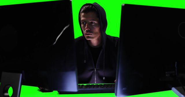 Hooded individual working on computers with two monitors and a visible green screen background. Ideal for use in articles about cybersecurity, hacking, technology risks, IT stories, and educational materials on internet security and cybercrime prevention.