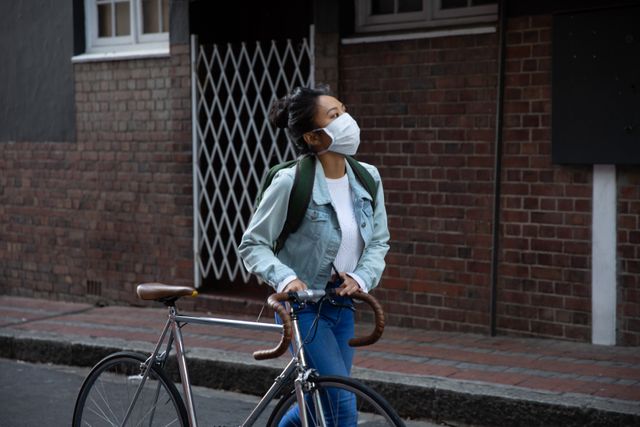 Biracial woman wearing a face mask against coronavirus, wheeling her bicycle in an urban setting. She is carrying a backpack and looking at buildings, suggesting a casual day out in the city. This image can be used for topics related to health and safety during the pandemic, urban lifestyle, transportation, and outdoor activities.