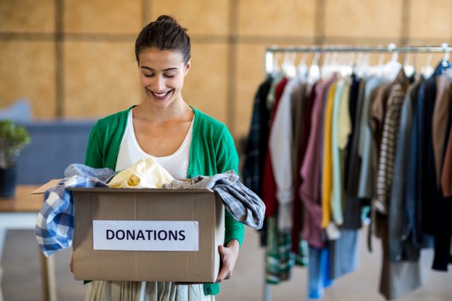 Woman holding a donation box filled with clothes in an office environment. Ideal for themes related to charity, community service, volunteer work, and support. Perfect for illustrating articles, blog posts, or advertisements promoting charitable activities or volunteer organizations.