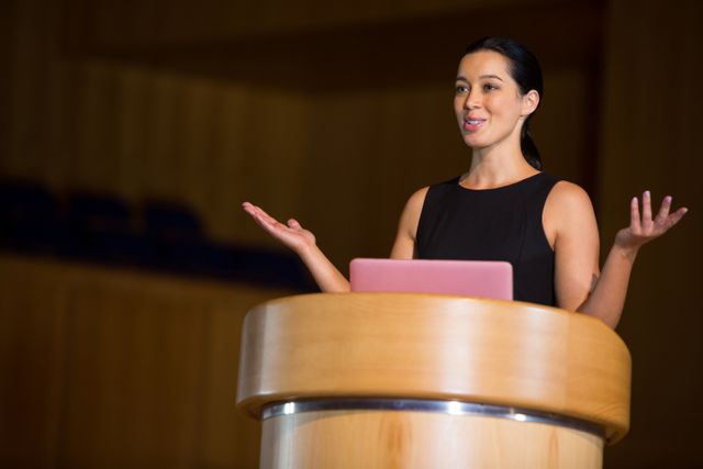 Female executive confidently giving a speech at a conference center, standing at a podium. Perfect for use in business articles, leadership presentations, public speaking workshops, corporate event promotions, and motivational speaker profiles.