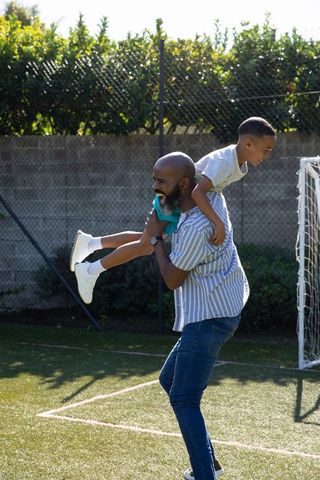 Father Carrying Child Piggyback on Backyard Soccer Pitch Joyfully