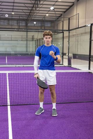 Young male padel player standing at net on purple indoor court preparing serve with racket and ball