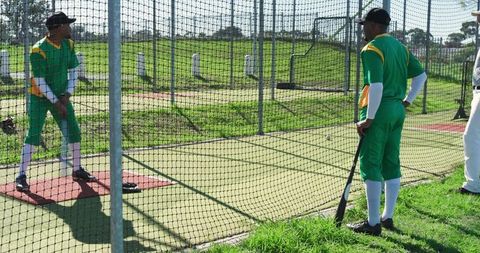 Cricket teammates practicing batting inside net enclosure during sunny outdoor training