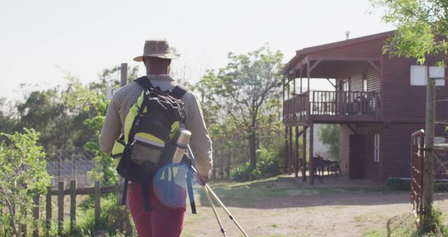 A hiker is approaching a rural wooden cabin, surrounded by greenery and trees. He carries a large backpack and uses walking sticks, embodying a spirit of adventure and outdoor exploring. Perfect for use in articles or advertisements related to travel, hiking, rural retreats, adventure tourism, and nature exploration.