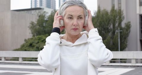 Senior woman adjusting headphones on urban rooftop wearing white hoodie, calm expression