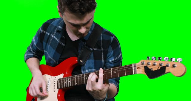 Young man wearing a plaid shirt playing an electric guitar against a green screen background. Ideal for usage in music videos, concert promotions, music instructional materials, or advertisements related to musical instruments and accessories.