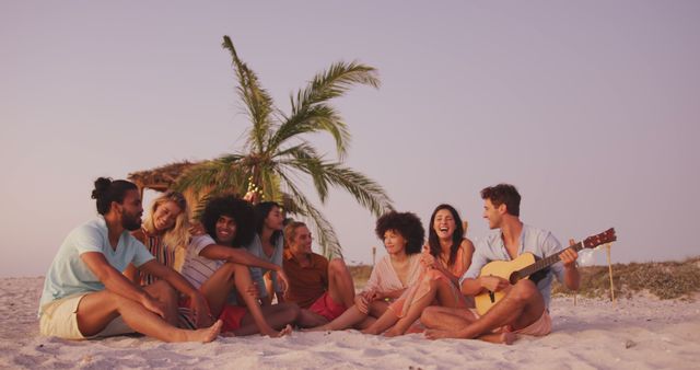 Happy diverse group of friends sitting on the beach playing guitar, relaxing and talking at sunset. Summer, free time, friendship, party, celebration and vacations.