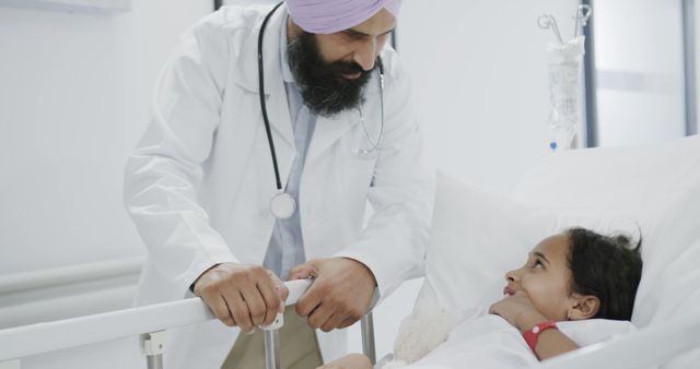 Doctor wearing a white coat talking to a young patient lying in a hospital bed. The doctor appears compassionate and attentive, engaging the child patient. Useful for healthcare campaigns, medical educational materials, and emphasizing doctor-patient relationships.