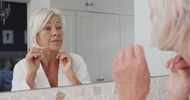 Elderly woman examining skin condition in bathroom mirror, wearing white bathrobe. Perfect for illustrating subjects related to senior health, skincare routines, beauty care for aging individuals, and self-awareness practices among the elderly.