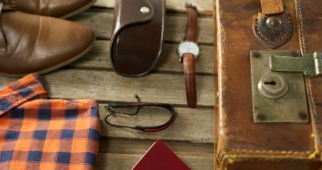 A collection of men's accessories and travel items are laid out on a wooden surface, including a plaid shirt, leather shoes, a watch, glasses, and a suitcase. These items suggest a sense of adventure or preparation for a journey, evoking a masculine and exploratory theme.