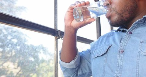 Man in Denim Drinking Water by Window for Refreshment and Hydration