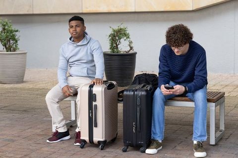 Diverse young men sitting on urban bench with suitcases waiting and using smartphone