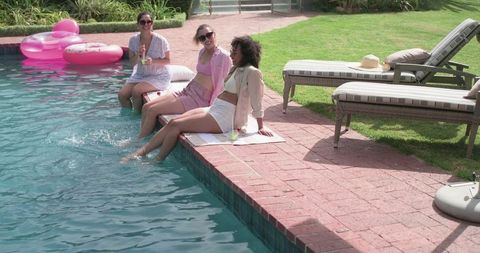 Three friends relaxing by backyard pool dipping feet and drinking
