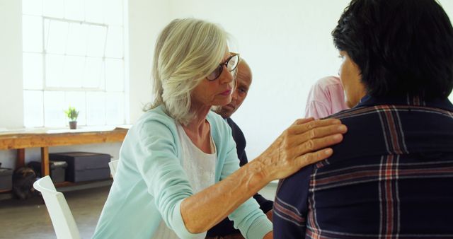 Elderly woman placing her hand on another person's shoulder expressing empathy and compassion in a support group setting. Both individuals and others are sitting indoors, indicating a group therapy session. This can be used for themes like mental health, community support, therapy sessions, counseling, emotional care, or senior citizens providing guidance.