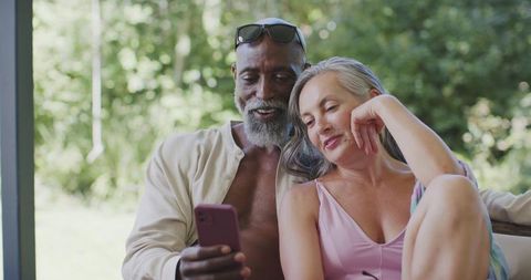 Middle-aged diverse couple relaxing on sunlit patio scrolling phone together, cozy moment