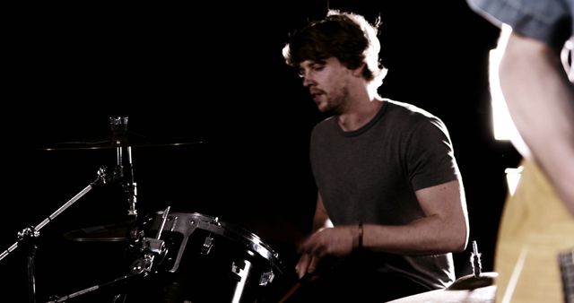 Drummer performing in a dimly lit music studio, focused on playing drums. Useful for content related to live music performances, musical talent, band practices, and artistic visuals of music production. Ideal for promoting musical events, illustrating music-related articles, or creating advertisements for music schools.