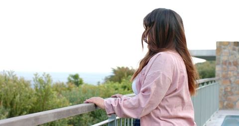Woman leaning on railing gazing at ocean from coastal terrace wearing pink jacket
