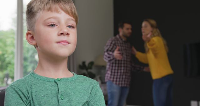 Young boy standing with a closed expression, while his parents argue in the background in a domestic setting. This stock photo can be used to depict themes related to family conflicts, parenting challenges, children's emotional responses, and the effects of arguments on children. Suitable for articles or projects about family counselling, child psychology, and social issues.