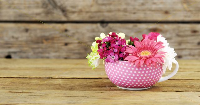 Bright and vibrant flowers arranged in a pink polka dot teacup on rustic wooden table perfect for spring-themed projects. Ideal for home decor inspiration, floral arrangements, greeting card designs, and nurturing a cozy, rustic ambiance.