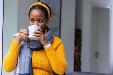 African american woman sipping coffee wearing mustard sweater, scarf and smartwatch by window
